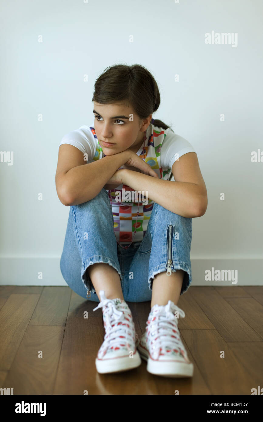 Teenage girl sitting on hardwood floor, hugging knees, looking away from camera Stock Photo