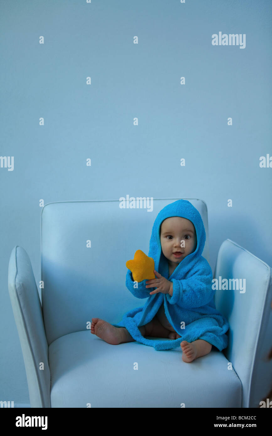 Baby boy wearing hooded bathrobe sitting in armchair, holding stuffed