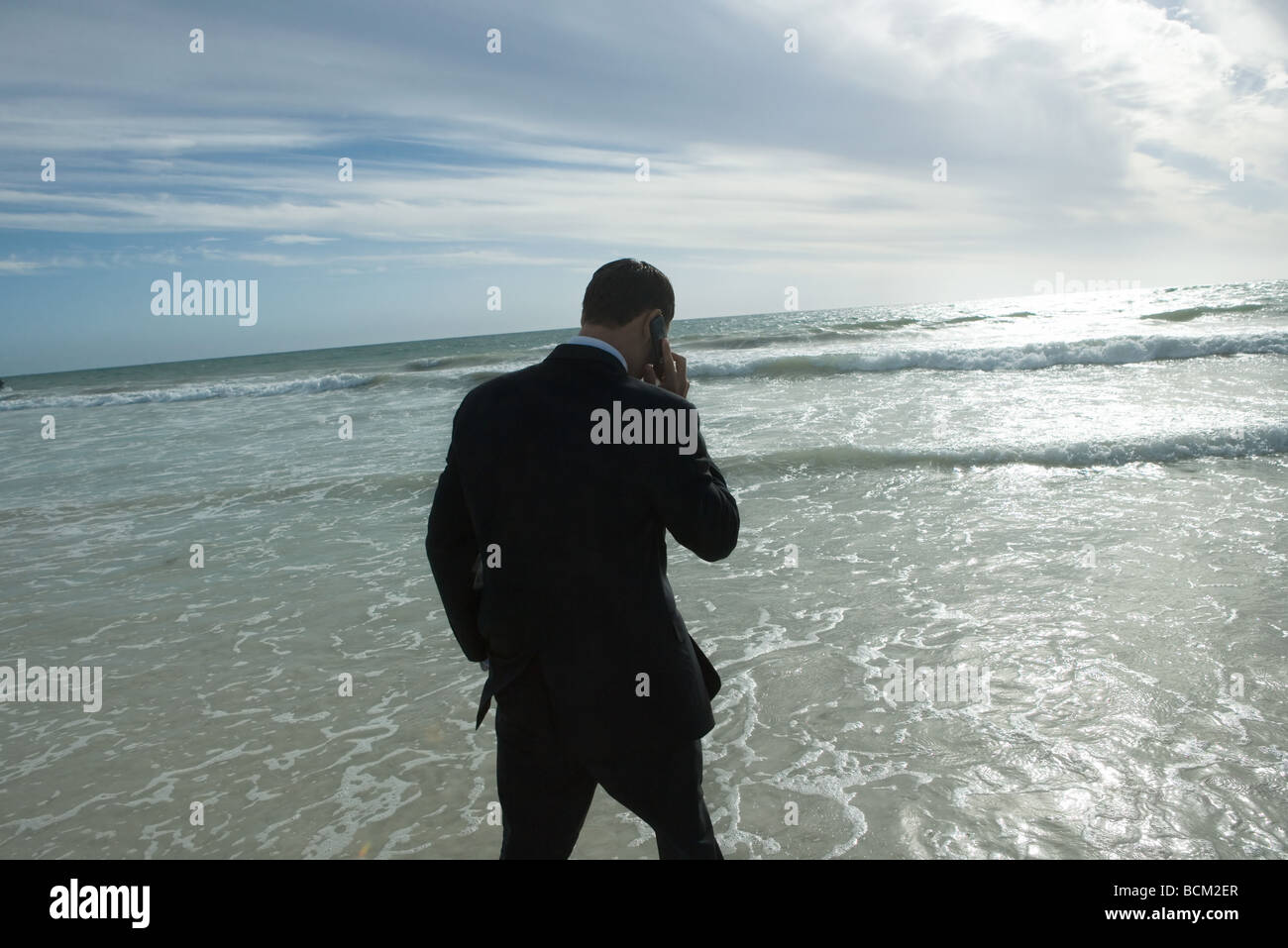 Businessman walking in surf at beach, talking on cell phone, rear view Stock Photo