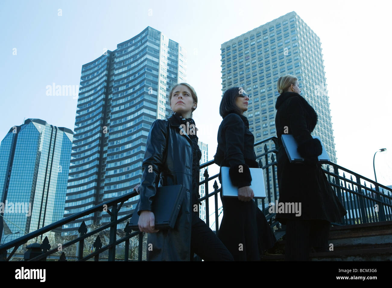 Three women dressed in black walking up steps, all carrying laptop computers, high rises in background Stock Photo