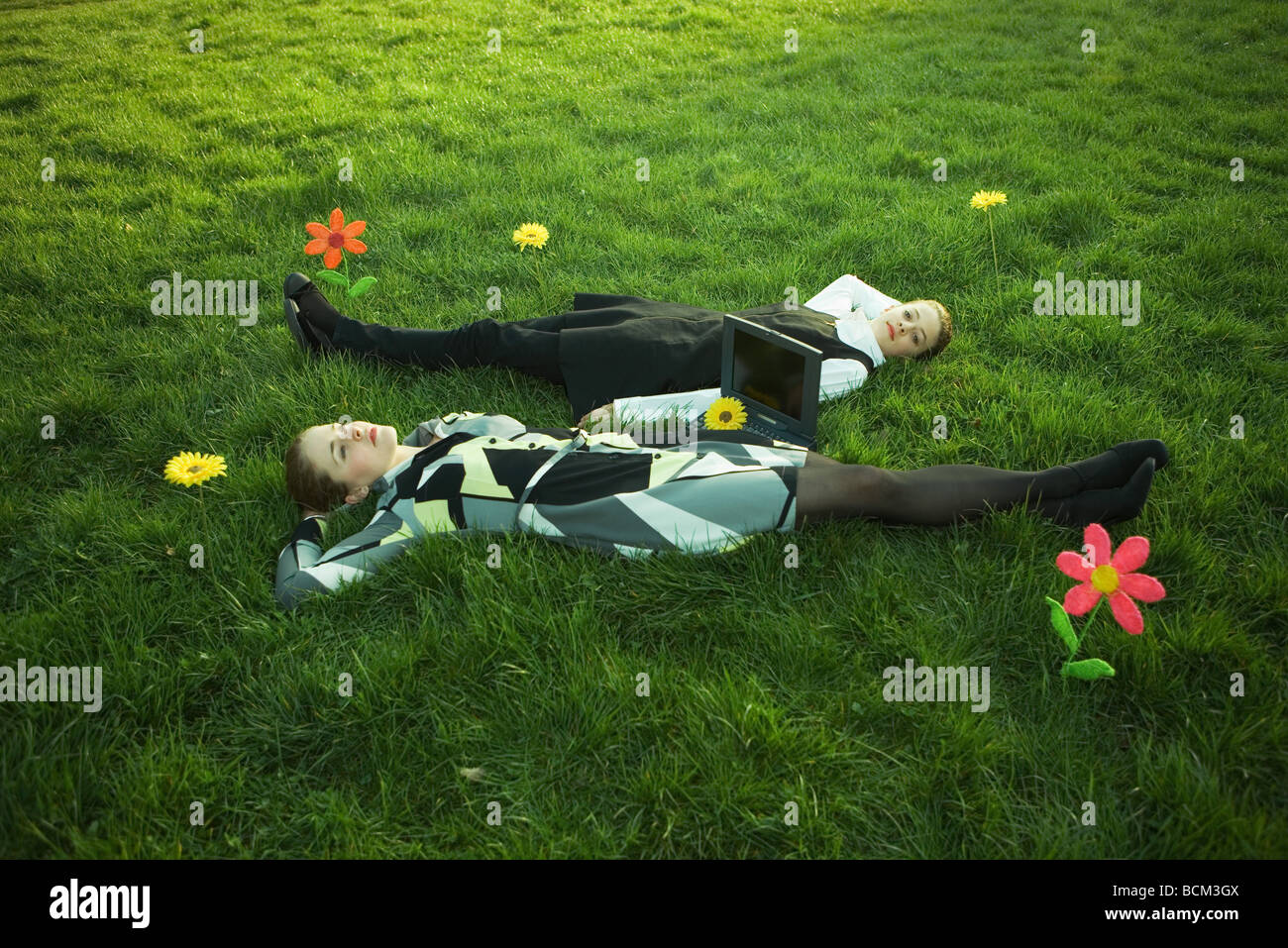 Two females lying in grassy field with flowers, laptop computer on the ground between them, one looking at camera Stock Photo