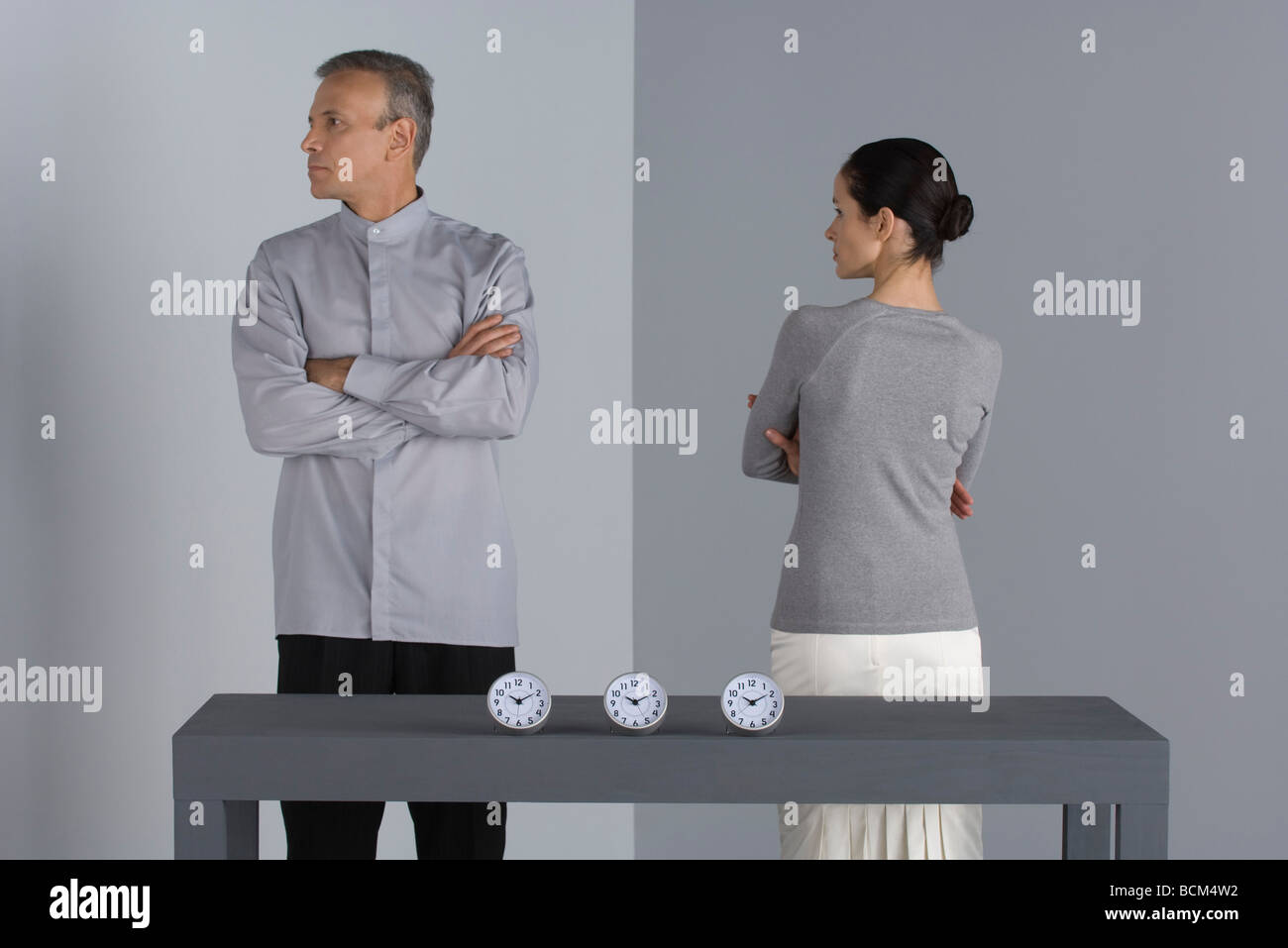 Couple standing with arms folded, backs turned to each other, clocks on table in front of them Stock Photo
