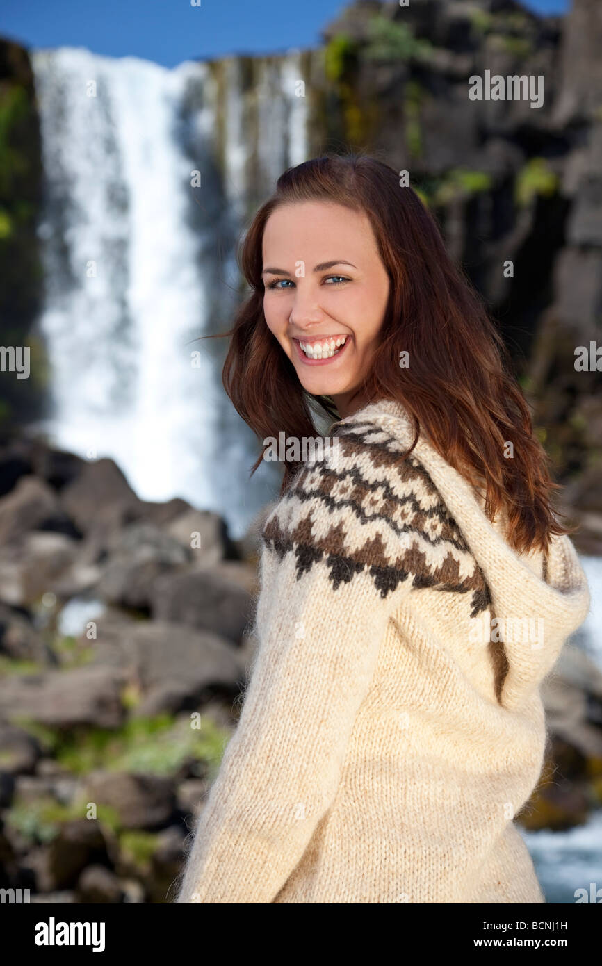 A beautiful Scandinavian woman wearing traditionally patterned knitwear standing and smiling in front of a mountain waterfall. Stock Photo