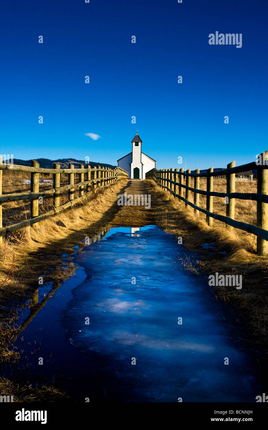 A white prairie church framed by blue sky, an icy pond reflecting a section of the steeple and rustic wooden fenceposts. Stock Photo