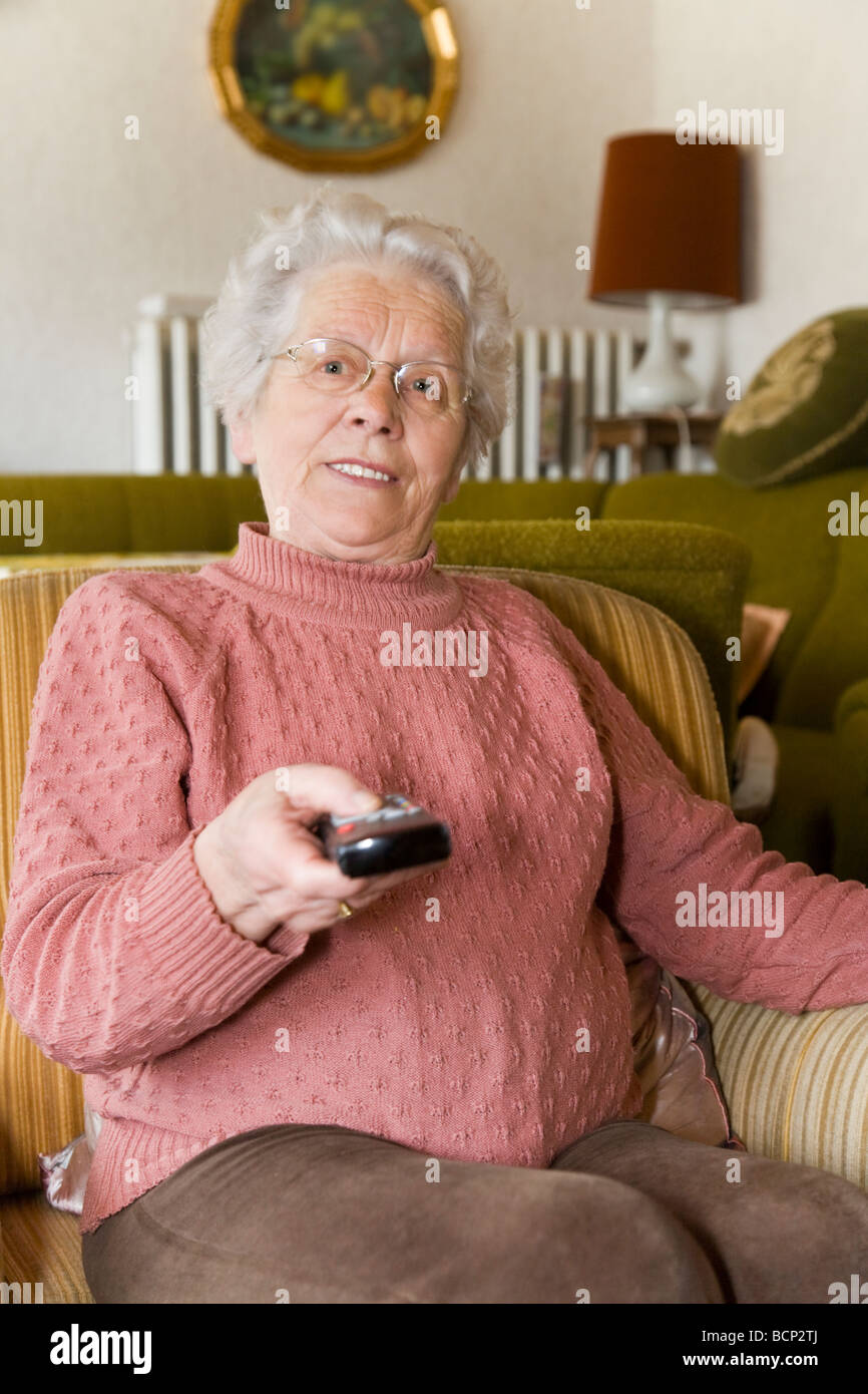 Frau in ihren Siebzigern sitzt im Wohnzimmer im Sessel mit einer Fernbedienung Stock Photo