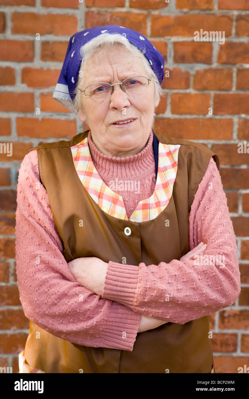 Frau in ihren Siebzigern steht mit Kopftuch Schürze und verschränkten Armen draußen vor einer Wand Stock Photo