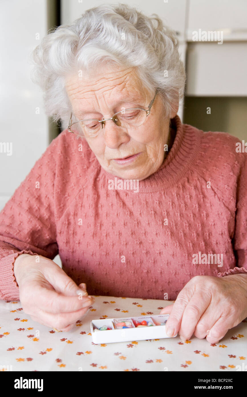 Frau in ihren Siebzigern sitzt am Küchentisch und schaut sich eine Tablette aus ihrer Pillendose an Stock Photo