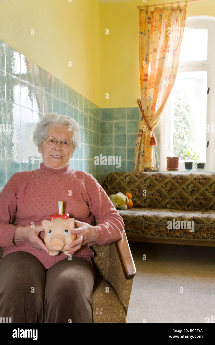 Frau in ihren Siebzigern sitzt auf einem Sessel und hält ein Sparschwein mit Geldscheinen Stock Photo