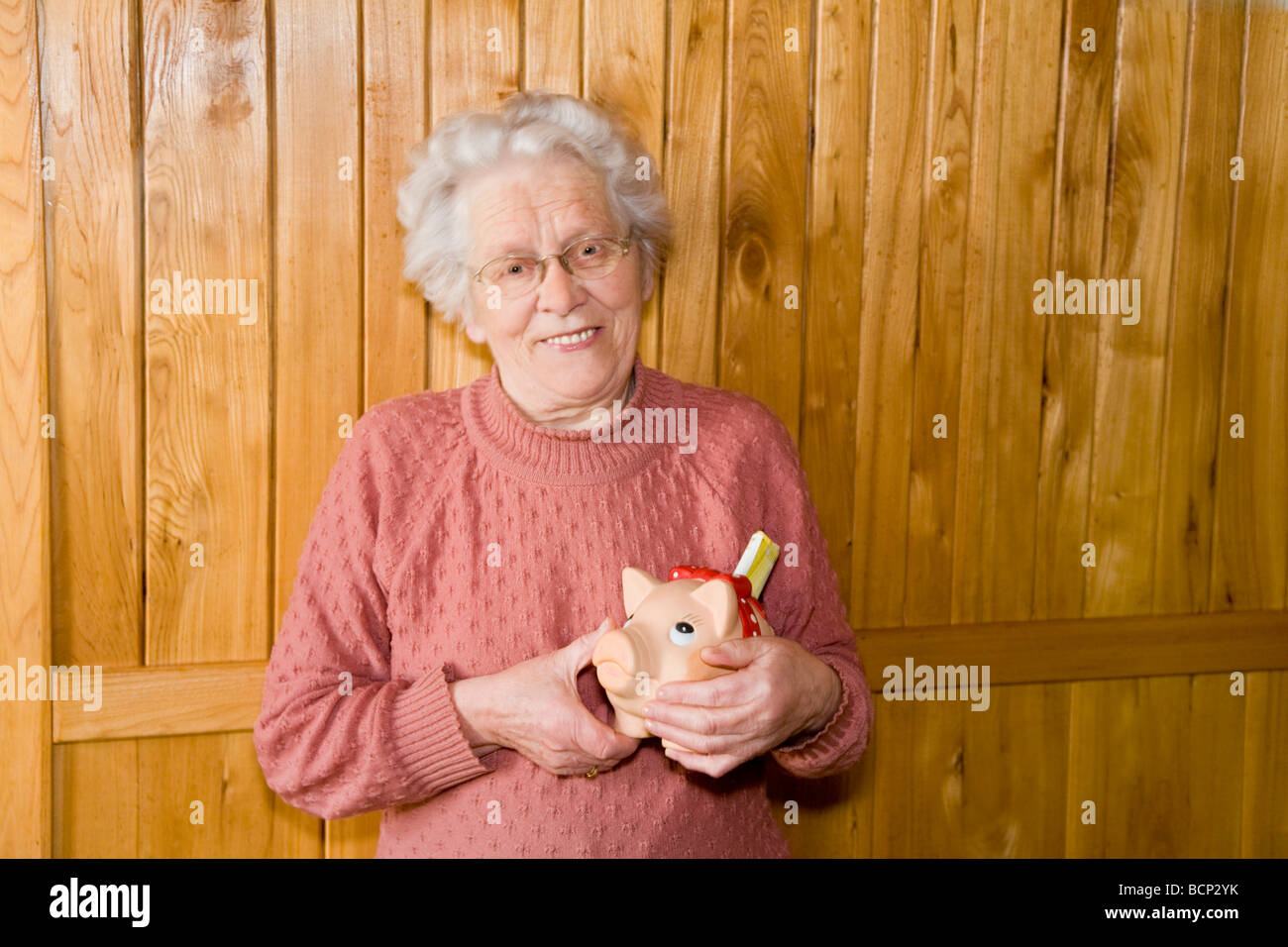 Frau in ihren Siebzigern steht vor einer Holzvertäfelung und hält lachend ein Sparschwein in den Händen Stock Photo