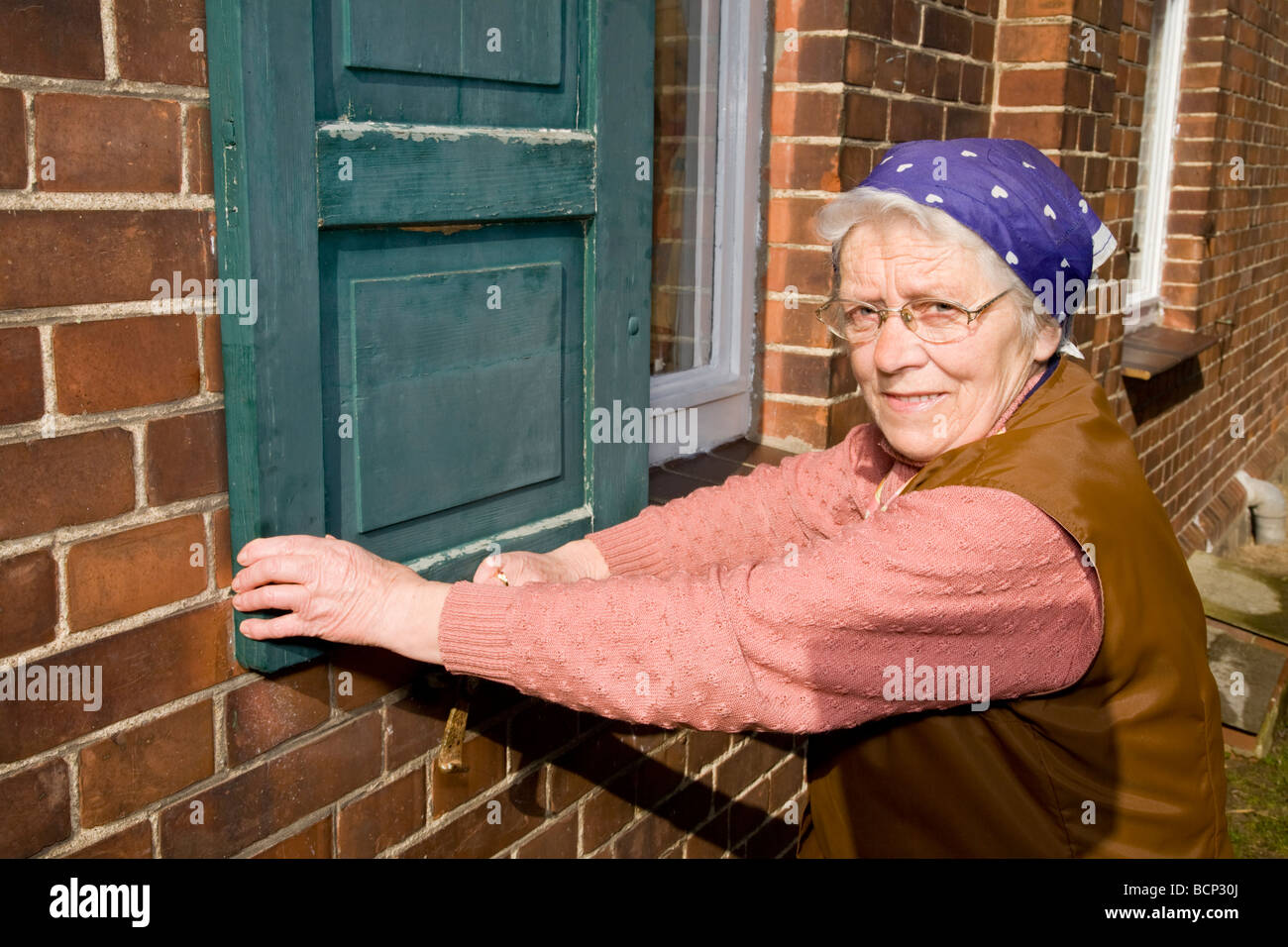 Frau in ihren Siebzigern mit Kopftuch schließt die Fensterläden eines Hauses Stock Photo