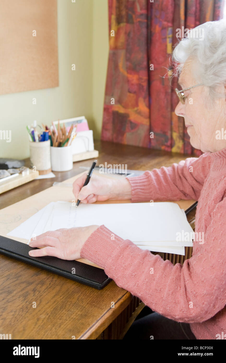 Frau in ihren Siebzigern sitzt am Schreibtisch und schreibt einen Brief Stock Photo