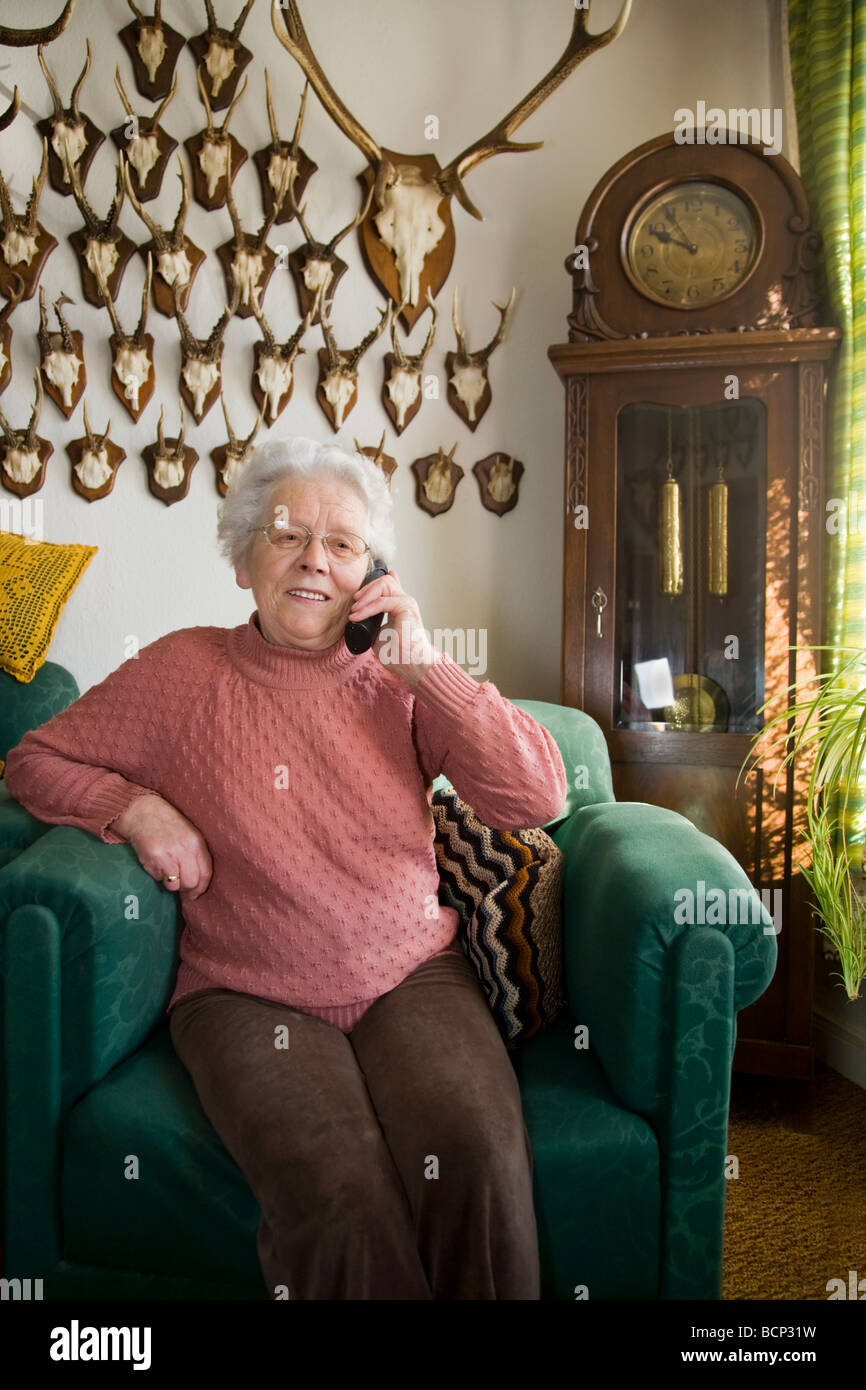 Frau in ihren Siebzigern sitzt in einem Sessel im Wohnzimmer und telefoniert mit einem schnurlosen Telefon Stock Photo