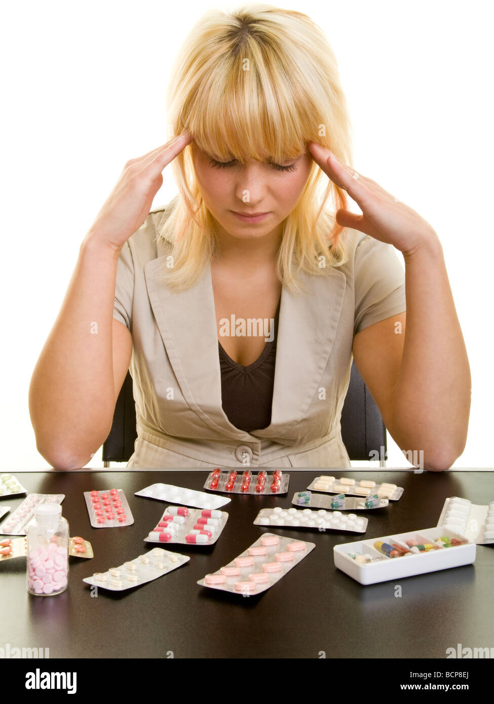 Junge Frau sitzt mit Kopfschmerzen vor einem Tisch mit vielen Arzneimitteln Stock Photo