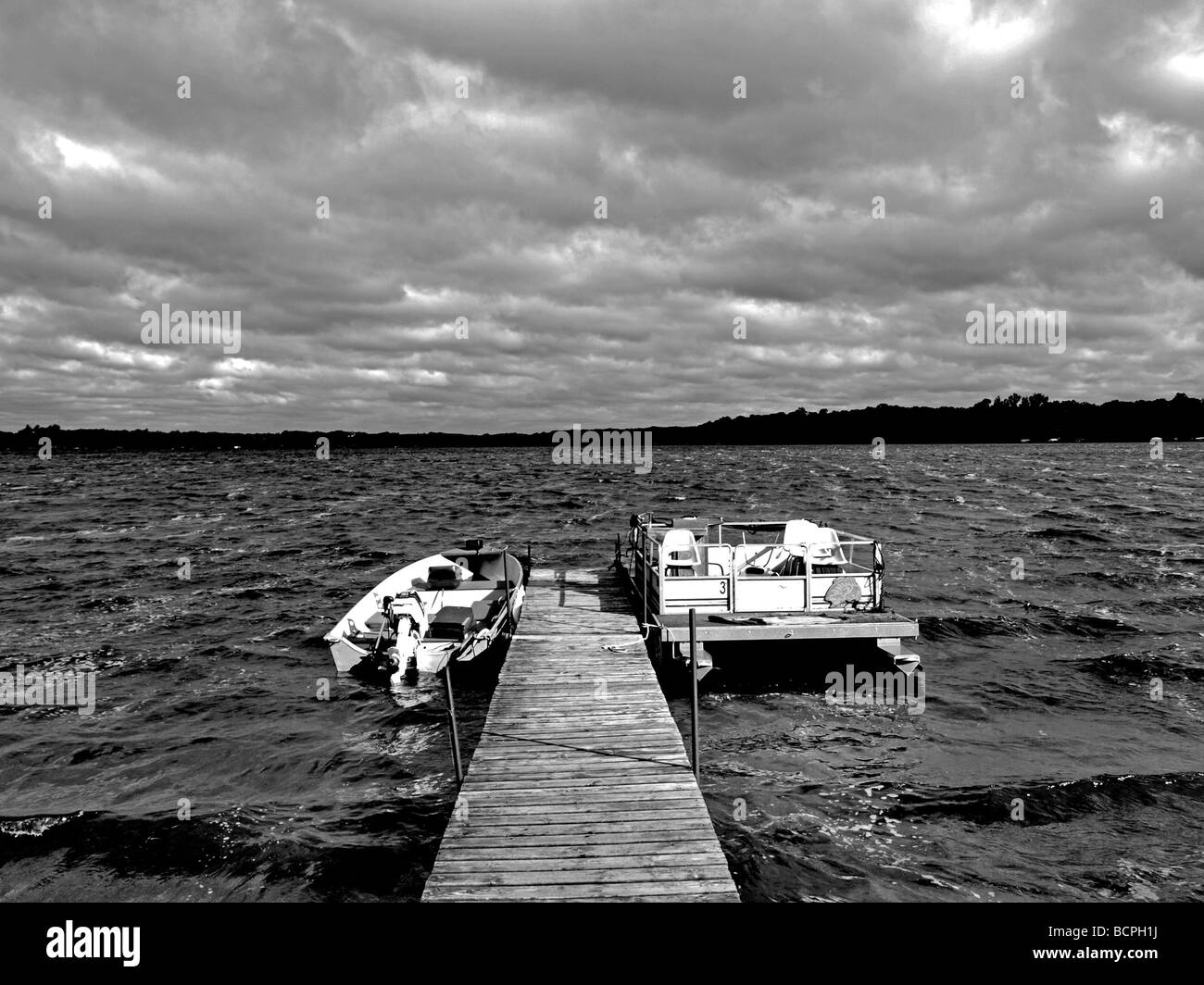 black and white image of two boats, motorboat with motor and the other a pontoon, tied to dock with gray clouds on lake Stock Photo