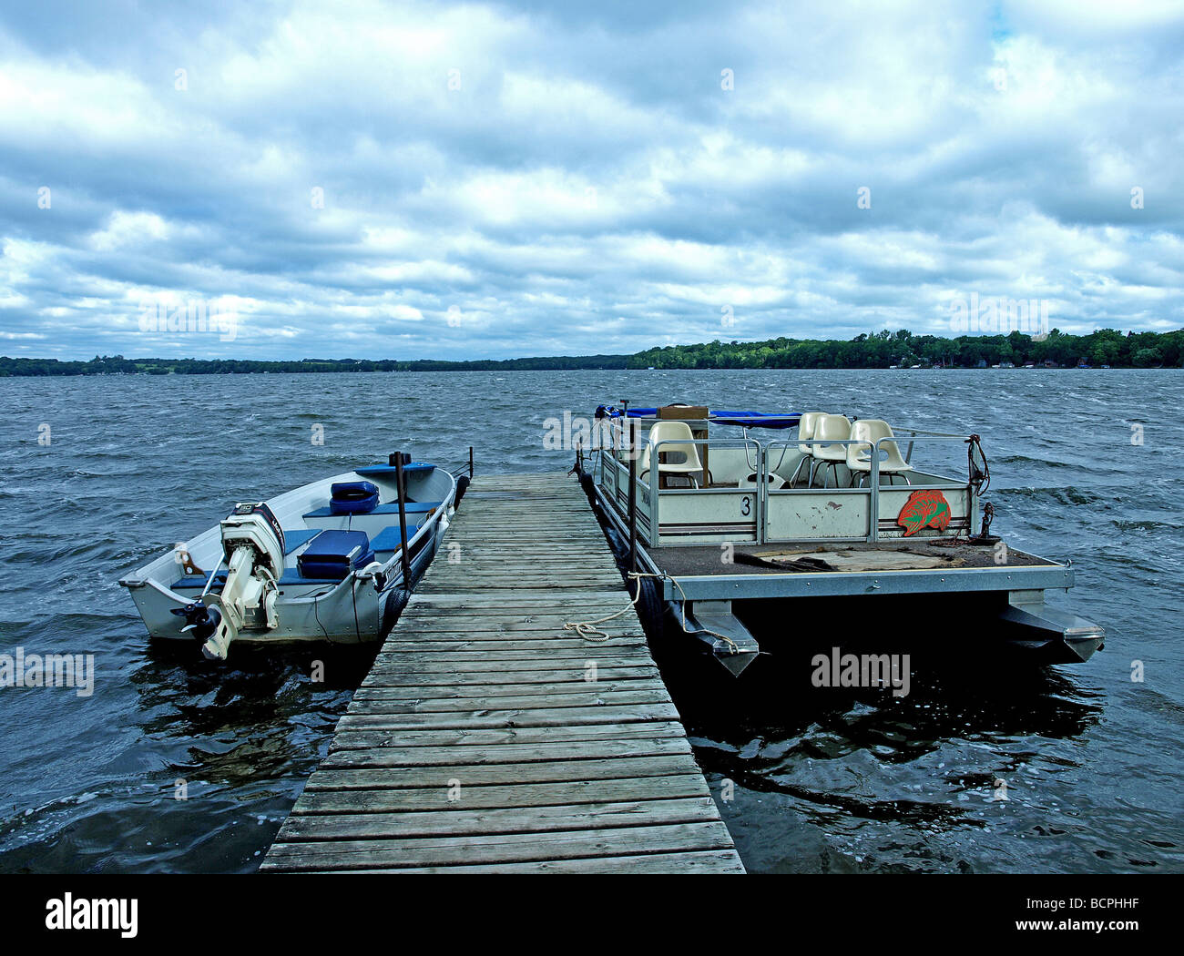 two boats tied to dock on wavy lake with cloudy skys and high waves, one boat is motorboat and the other is pontoon with chairs Stock Photo