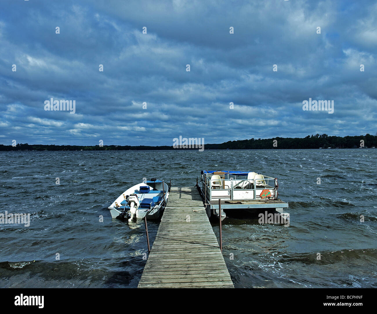 two boats tied to dock on wavy lake with cloudy skys and high waves, one boat is motorboat and the other is pontoon Stock Photo