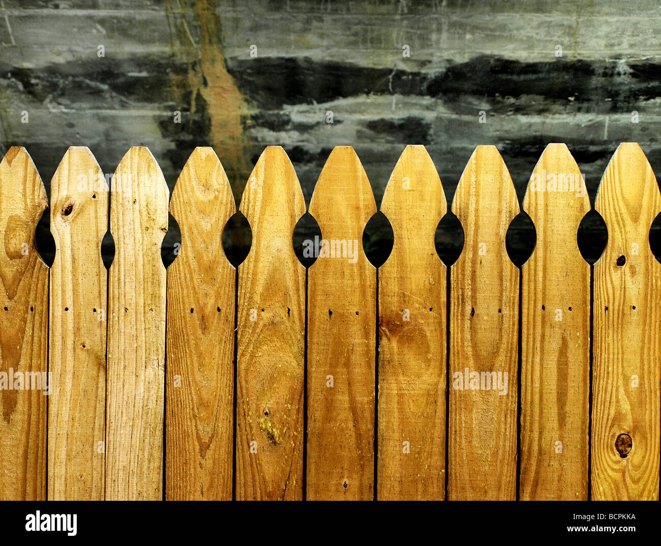 brown picket fence of wood with rounded tops in front of dark patterned cement wall Stock Photo