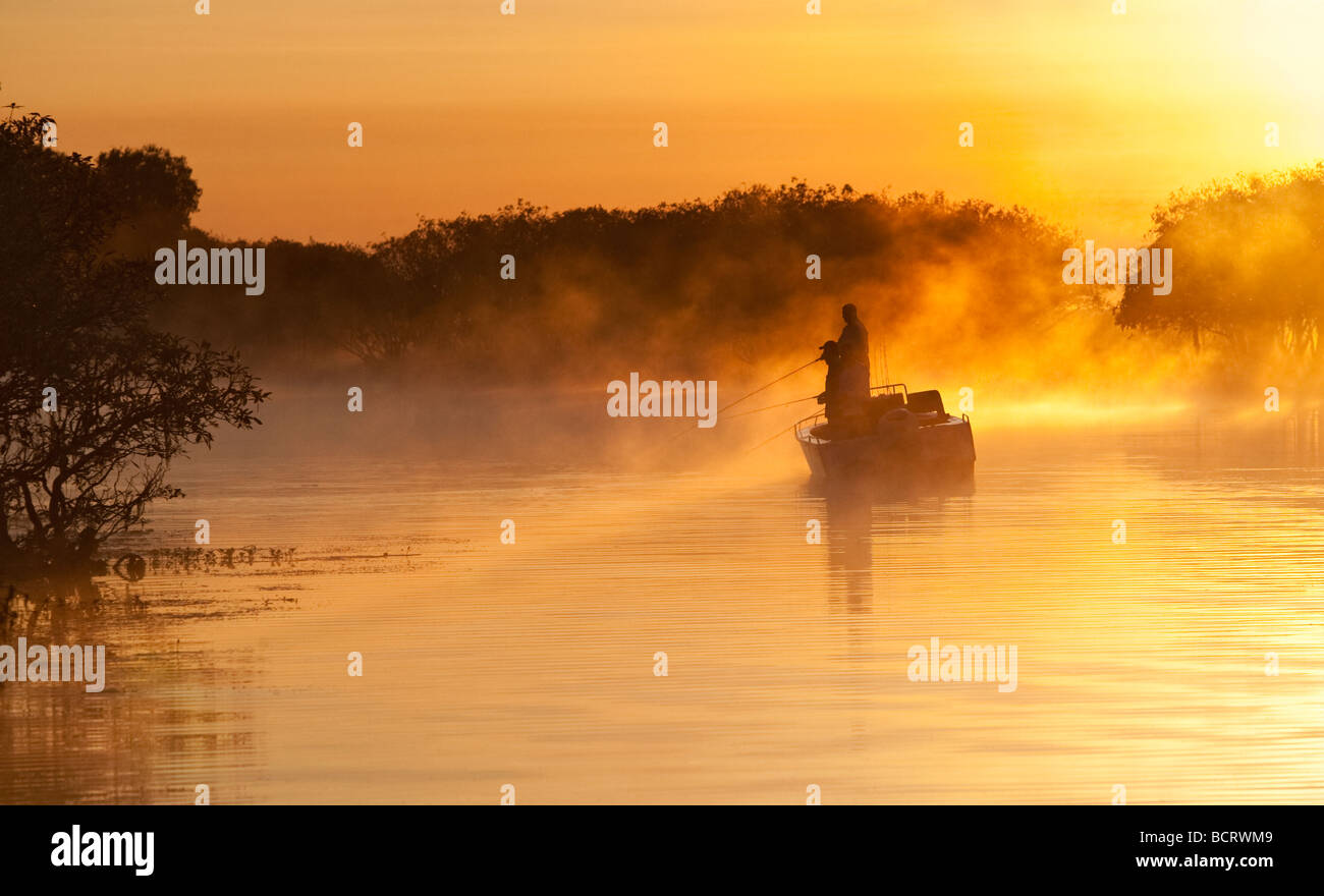 The sun rising behind a fishing boat while an early morning mist drifts over Yellow Water billabong in Kakadu National Park, Aus Stock Photo