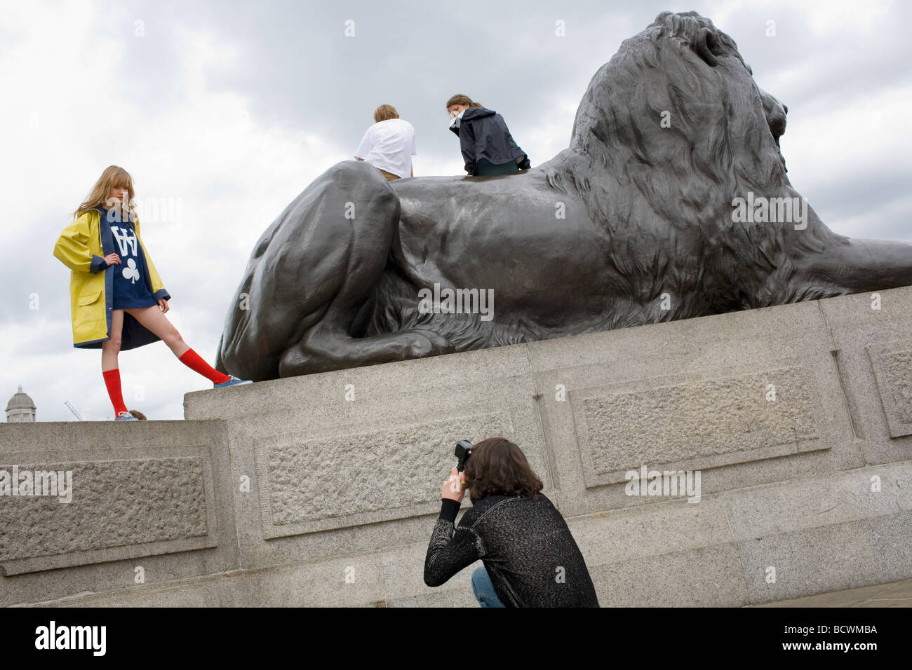 A young man photographs a pretty blonde girl in a yellow coat and red knee high socks posing by the lions in Trafalgar Square Stock Photo