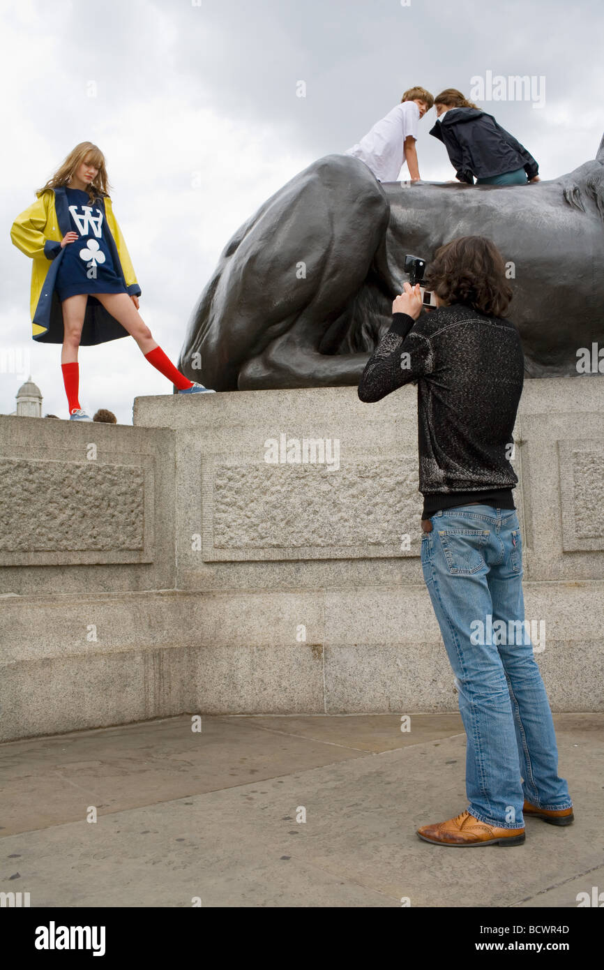 A young man photographs a pretty blonde girl in a yellow coat and red knee high socks posing by the lions in Trafalgar Square Stock Photo