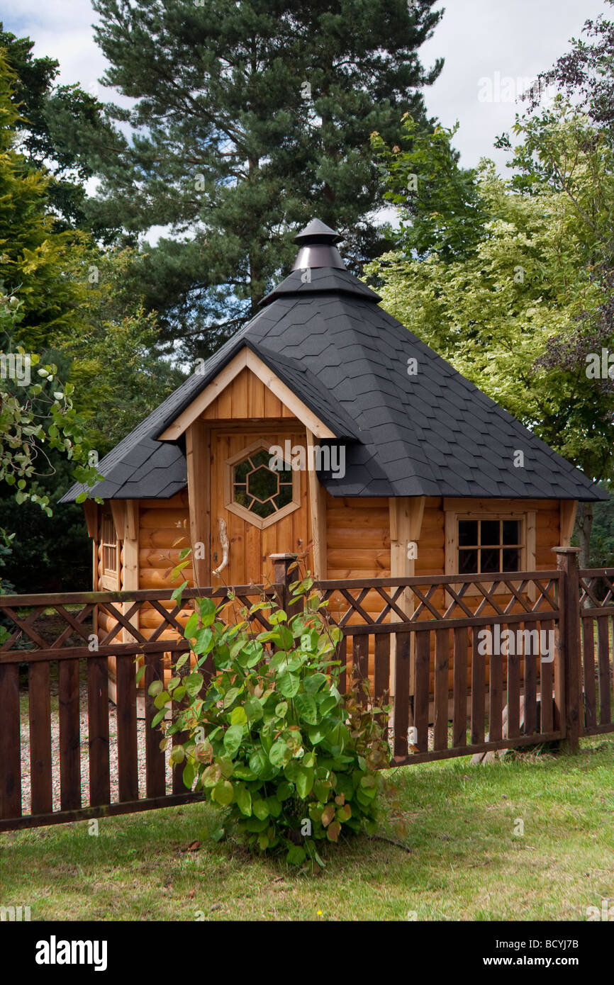 Wooden hexagonal tiled Garden shed with chimney, felt tiled roof and boundary fence; Deeside Log Cabins, Aberdeenshire, Scotland, UK Stock Photo