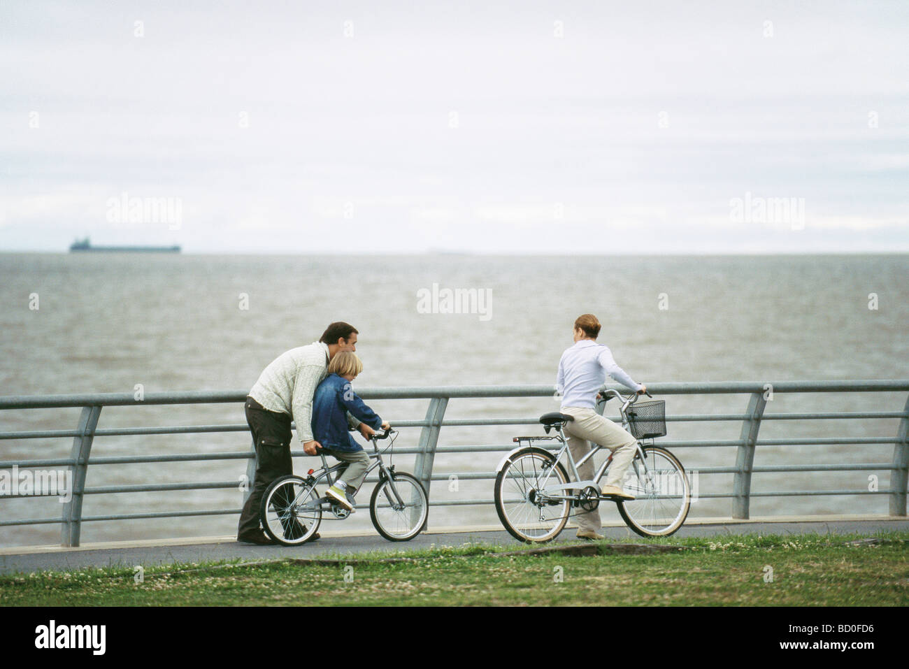 Father helping son learn to ride bicycle, mother watching Stock Photo