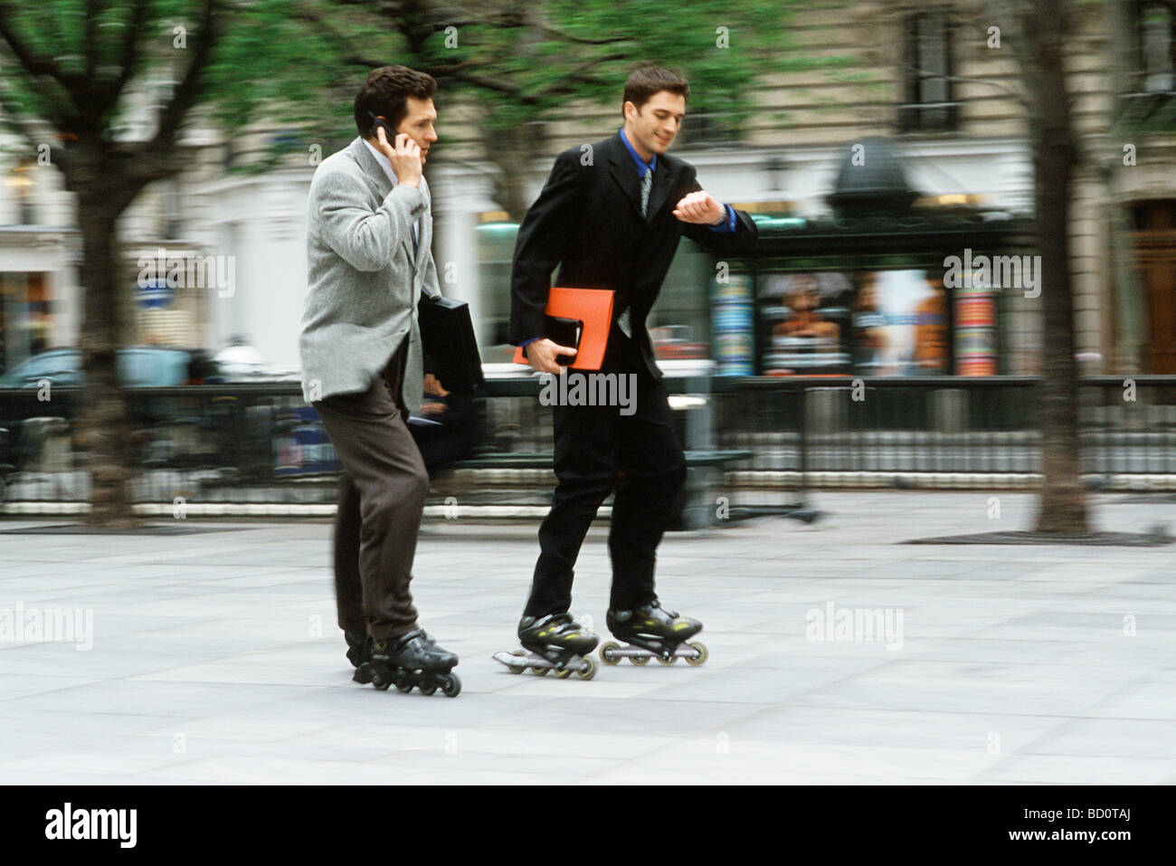Men in business attire rollerskating together along sidewalk, one phoning, the other looking at watch Stock Photo