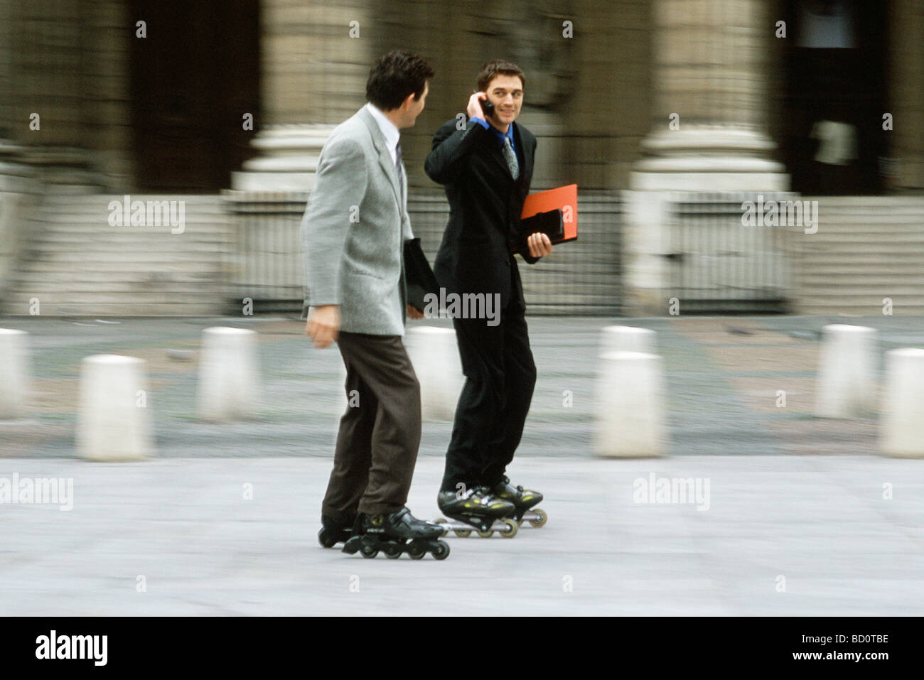 Men in business attire inline skating together along sidewalk, one phoning Stock Photo