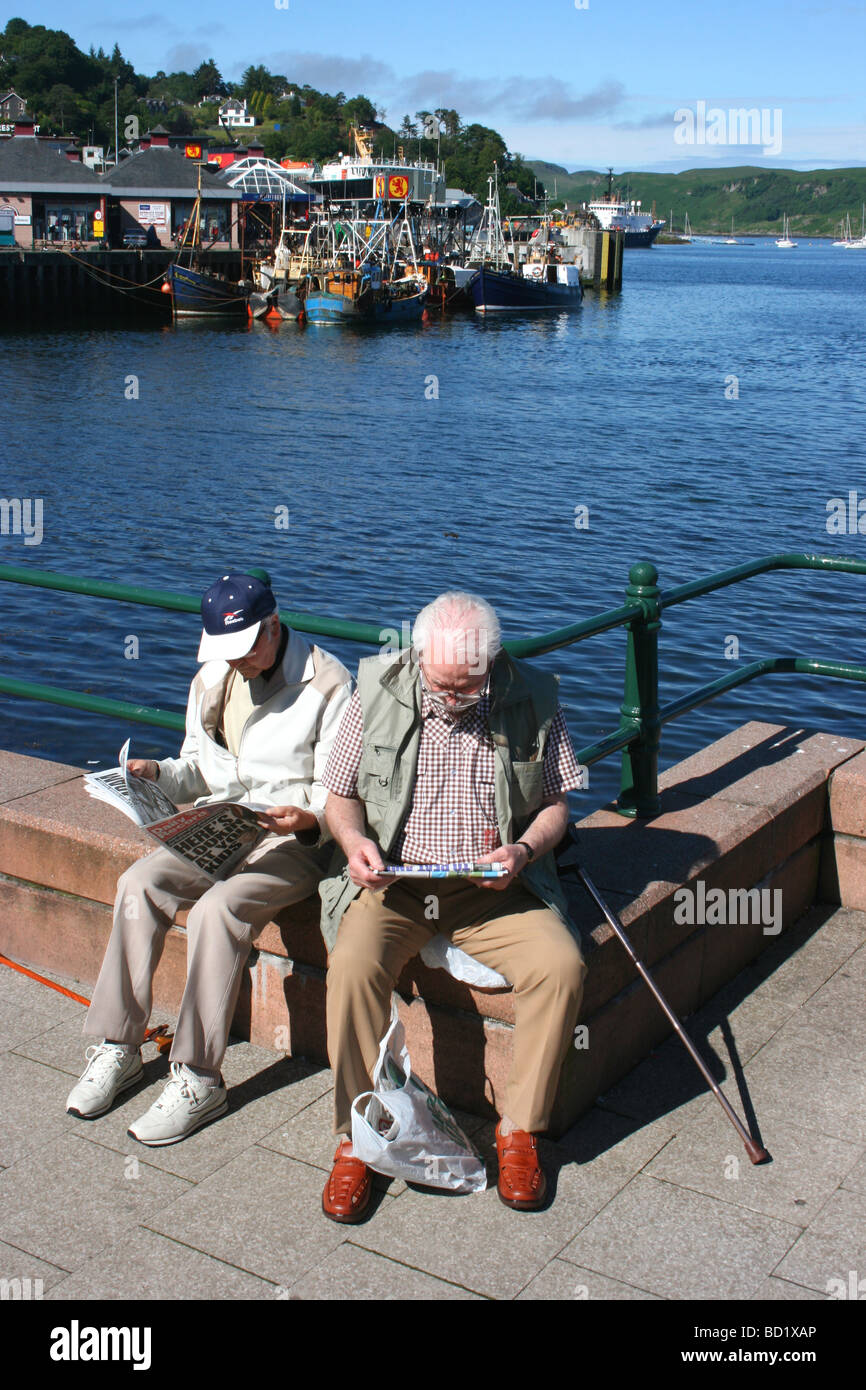 two old men relaxing, reading the morning newspapers on the promenade at Oban harbour, Argyll and Bute, West coast of Scotland Stock Photo