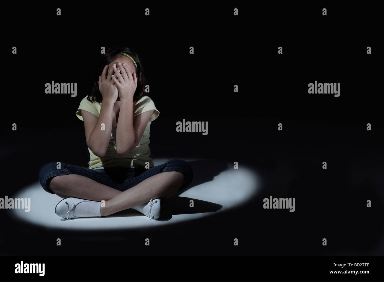 Young girl with hands covering her face sitting in spot light with dark background Stock Photo