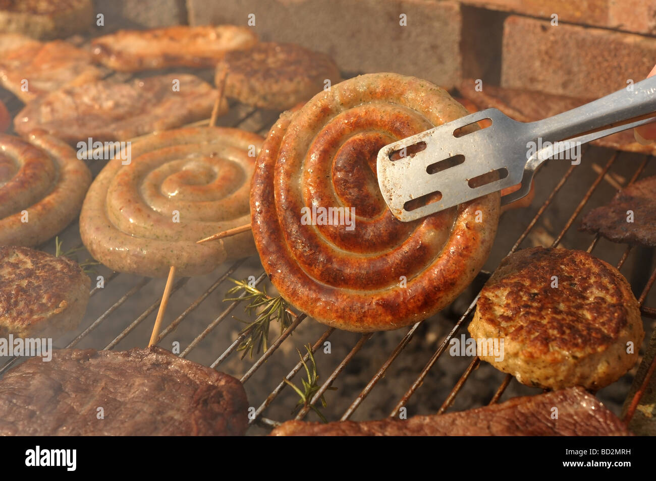 Smokey charcoal meat barbecue focus on tongs turning over a spiral Cumberland sausage Stock Photo