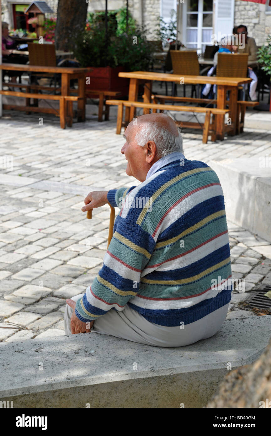elderly retired old man sitting relaxing looking observing resting on walking stick in the shade on bright sunny day in France Stock Photo