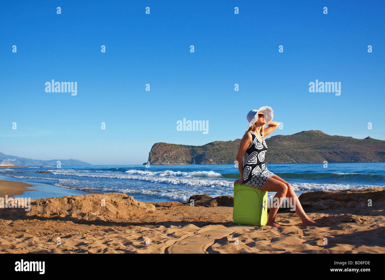 Young blond Caucasian woman wearing white straw hat and sunglasses sitting on green suitcase on a beach Stock Photo