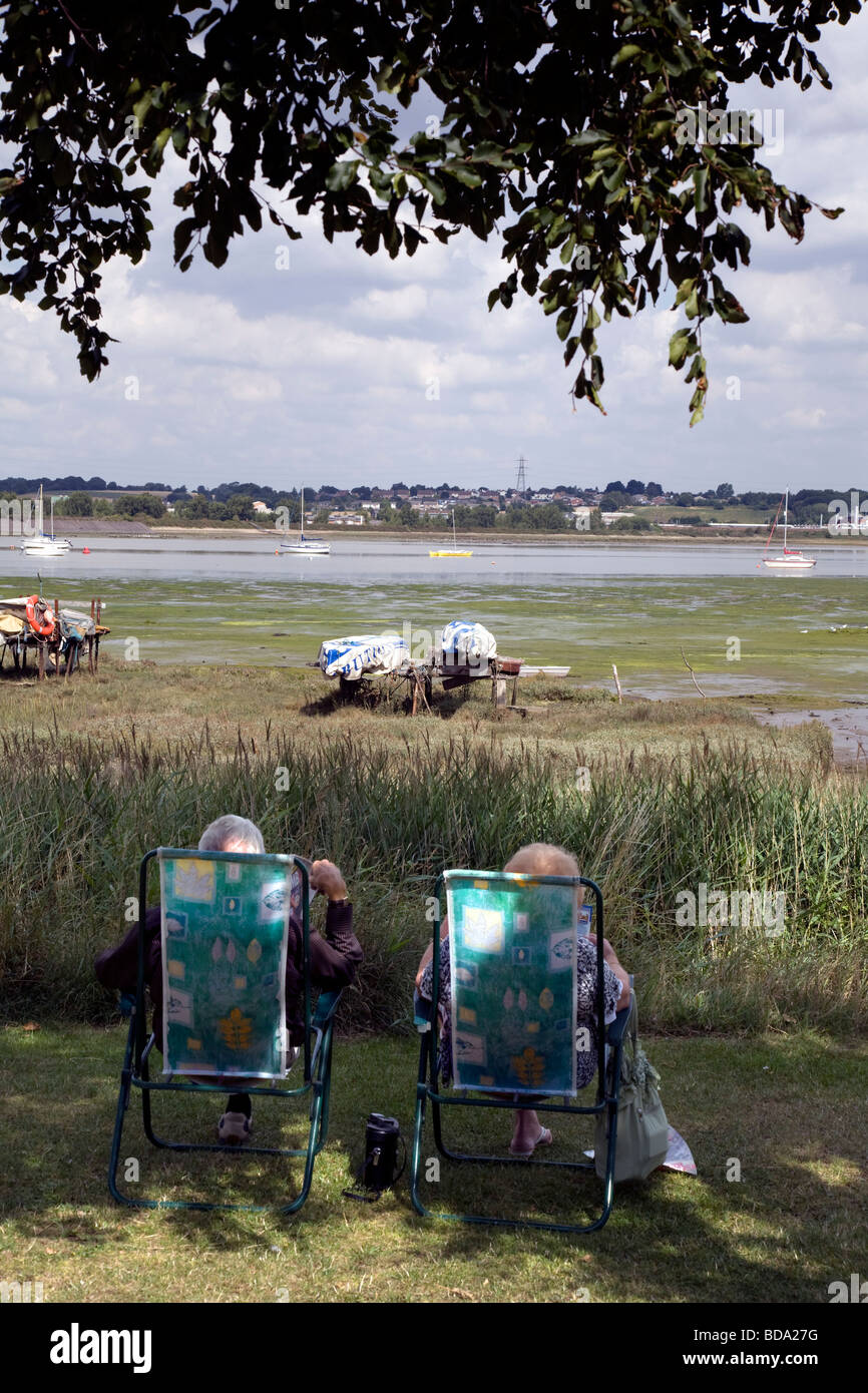 TWO ELDERLY PEOPLE SIT IN DECK CHAIRS BY THE RIVER STOUR, READING AT MANNINGTREE, BRITAIN'S SMALLEST TOWN Stock Photo