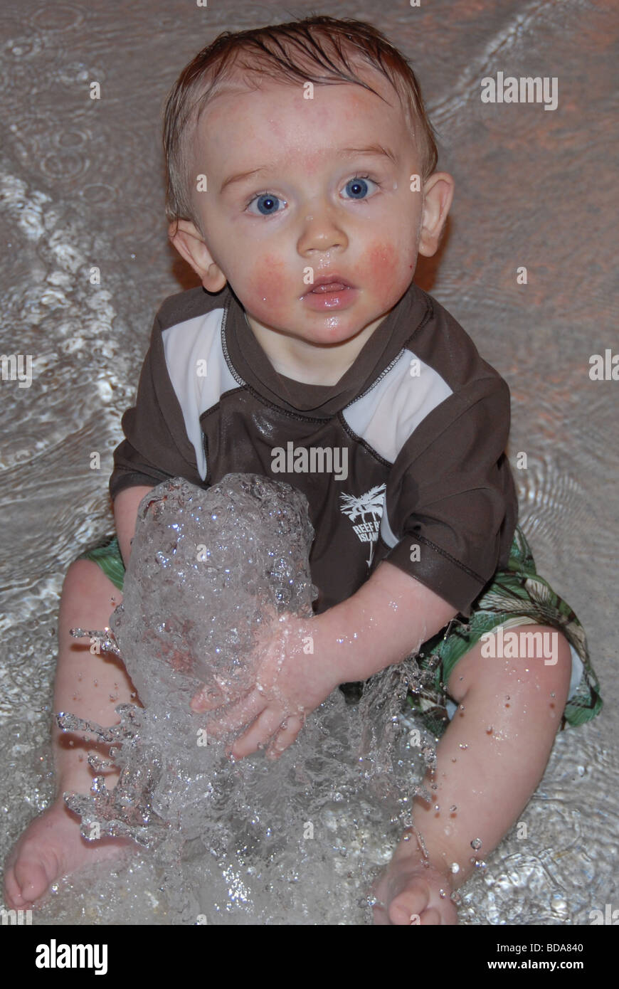 A baby boy sits in the water at a water park and plays with a small fountain. Stock Photo