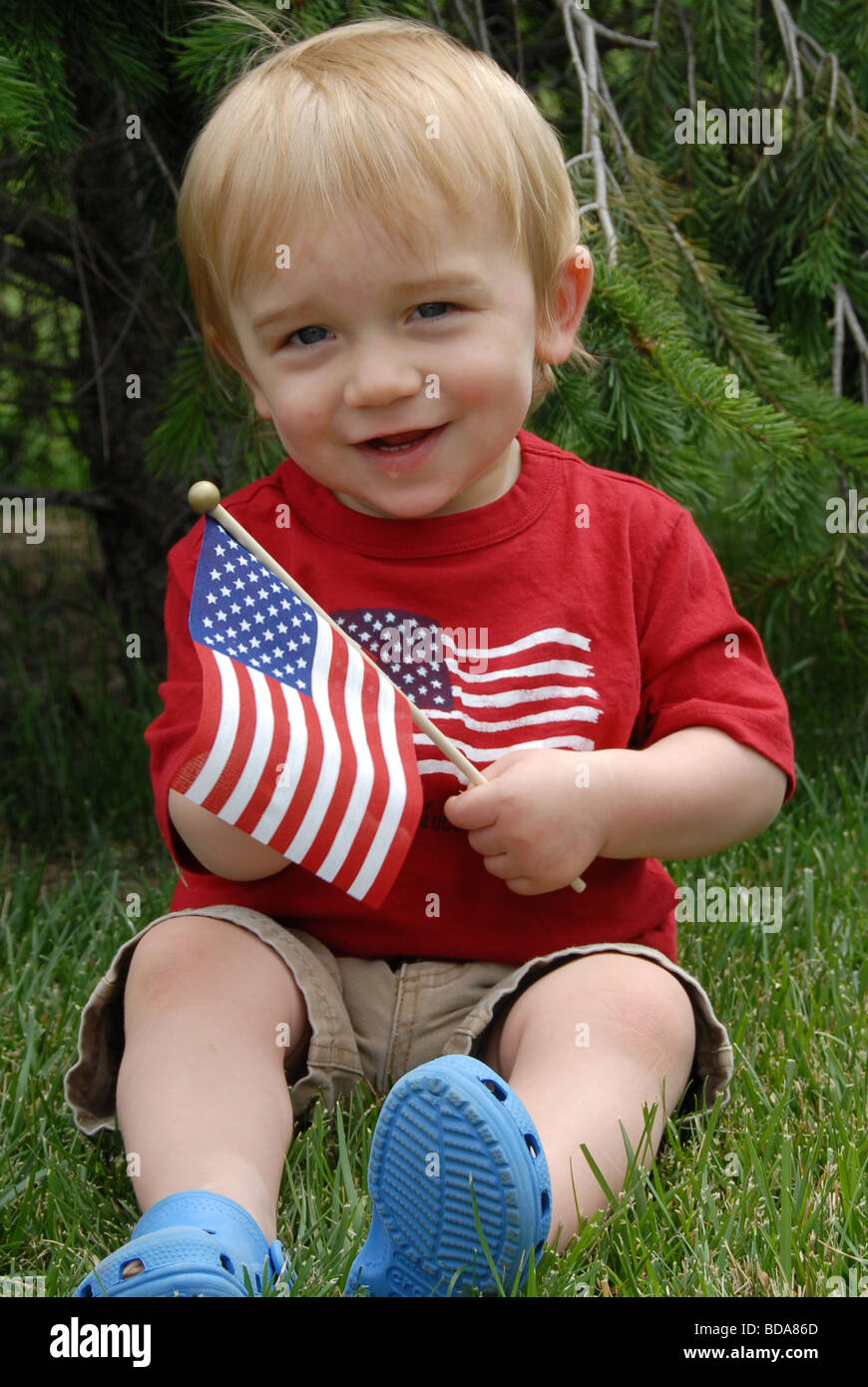 A baby boy sits in the grass waving an American flag on the Fourth of July wearing red white and blue clothes. Stock Photo