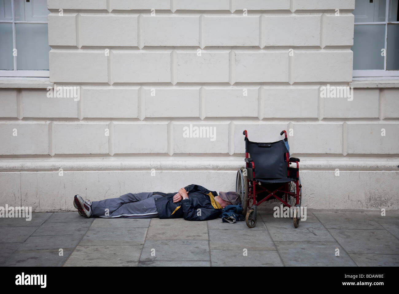 A homeless man lays next to his wheel chair sleeping perhaps passed out on the pavement, London Stock Photo