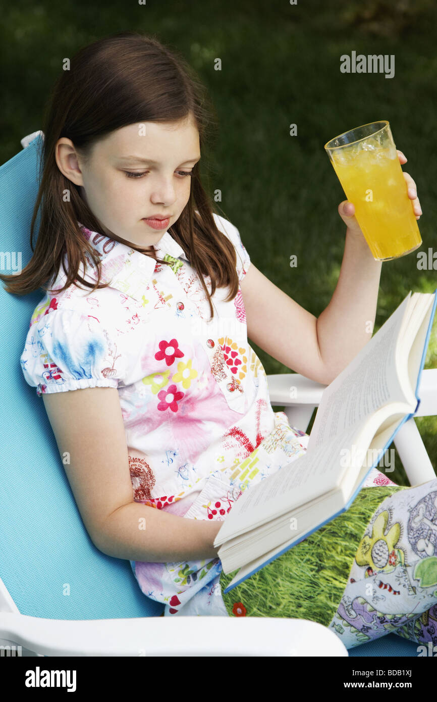 Girl holding a glass of juice and reading a book on a chair Stock Photo