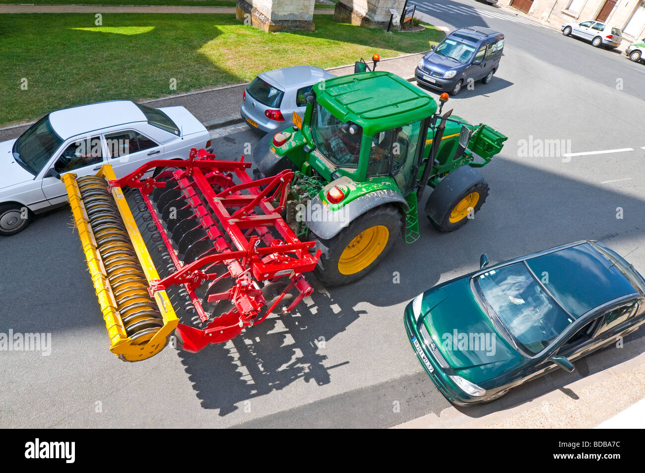 John Deere tractor & Terradisc harrow negotiating parked cars in town street - Indre-et-Loire, France. Stock Photo