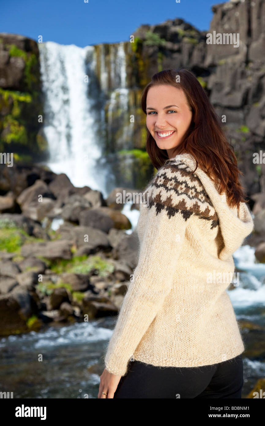 A beautiful Scandinavian woman wearing traditionally patterned knitwear smiling in front of a mountain waterfall Stock Photo