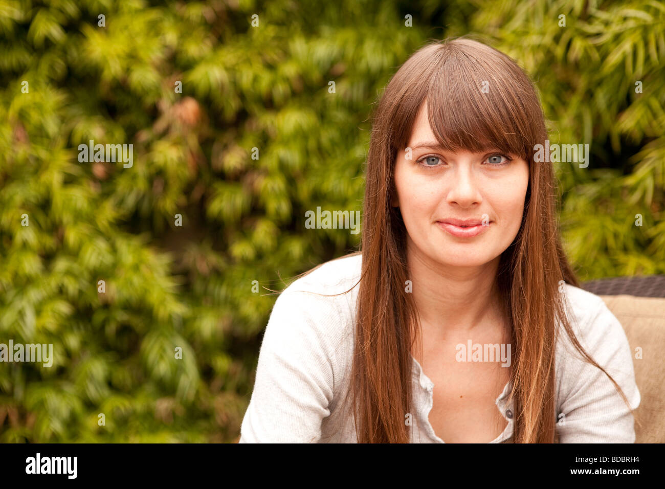 Upper body portrait of a woman with blue eyes and long brown hair sitting outside in-front of foliage. Stock Photo