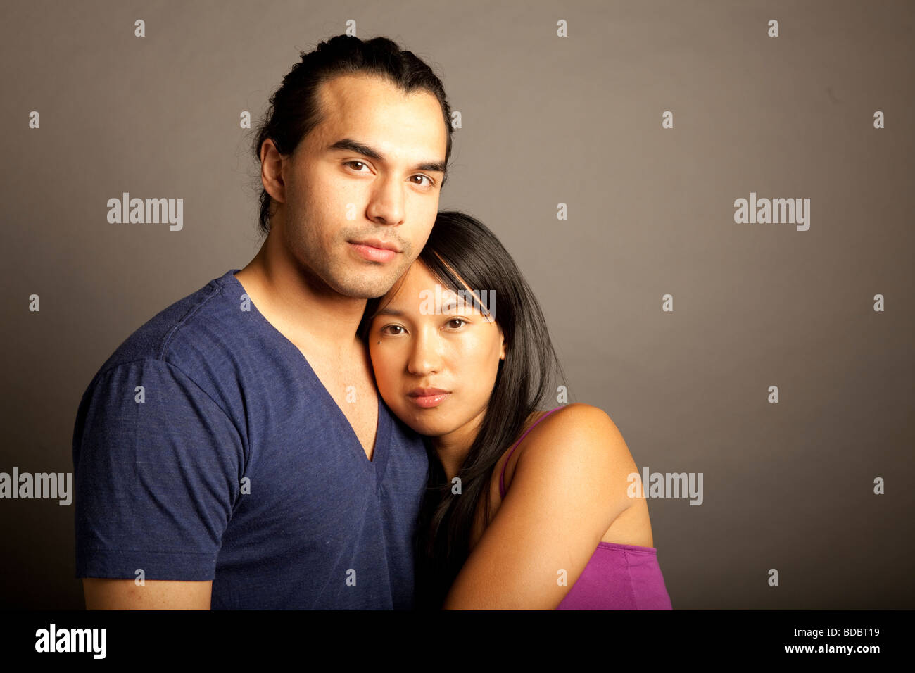 Portrait of Filipino female and ethnic male couple holding each other against gray background.  Looking at camera. Stock Photo