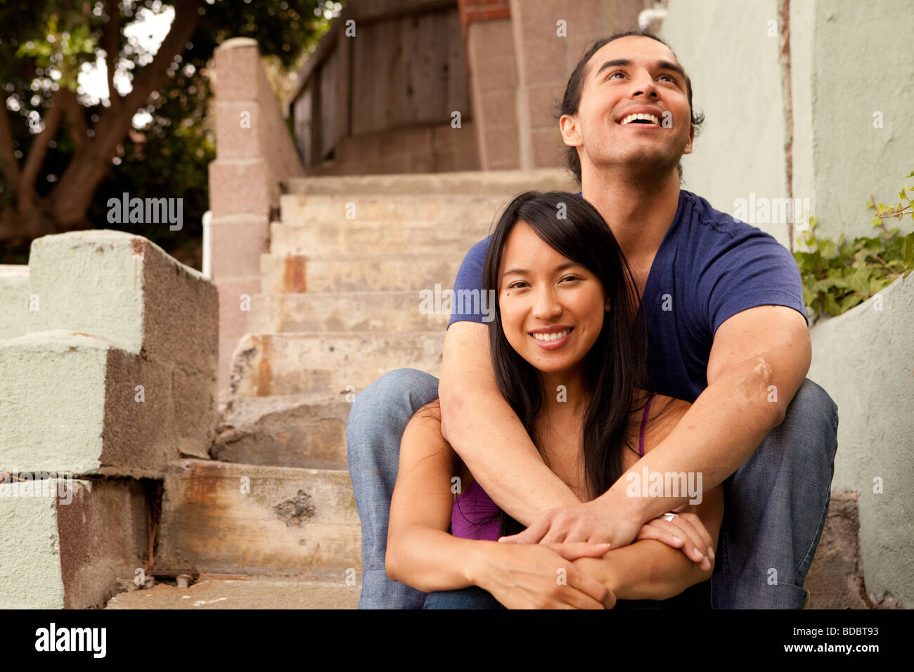 Male and female couple sitting on concrete steps, his arms are around her.  Looking at camera.  Woman is Filipino. Stock Photo