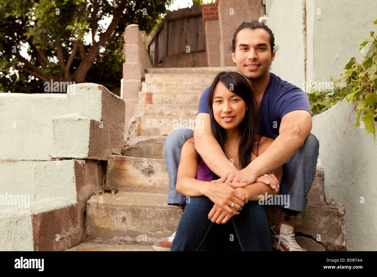 Male and female couple sitting on concrete steps, his arms are around her.  Looking at camera.  Woman is Filipino. Stock Photo