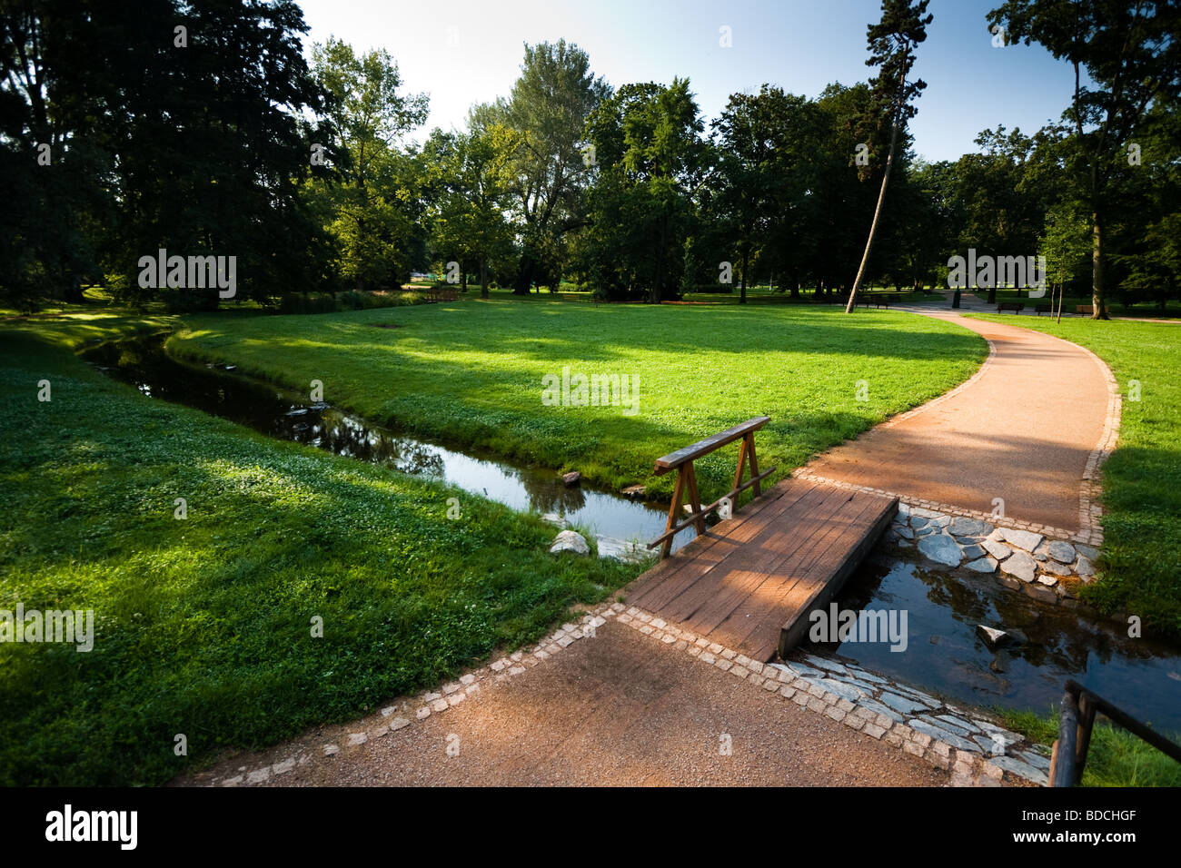 Summer morning in a park with road going across a small river via bridge. Stock Photo