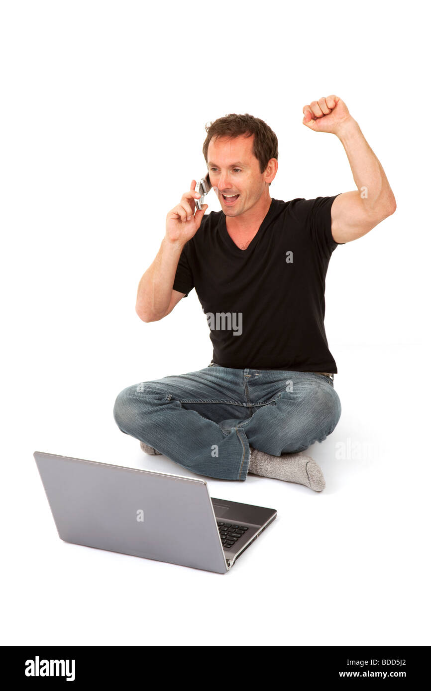 Happy middle aged man on mobile phone in front of laptop celebrating/punching air, on a white background Stock Photo