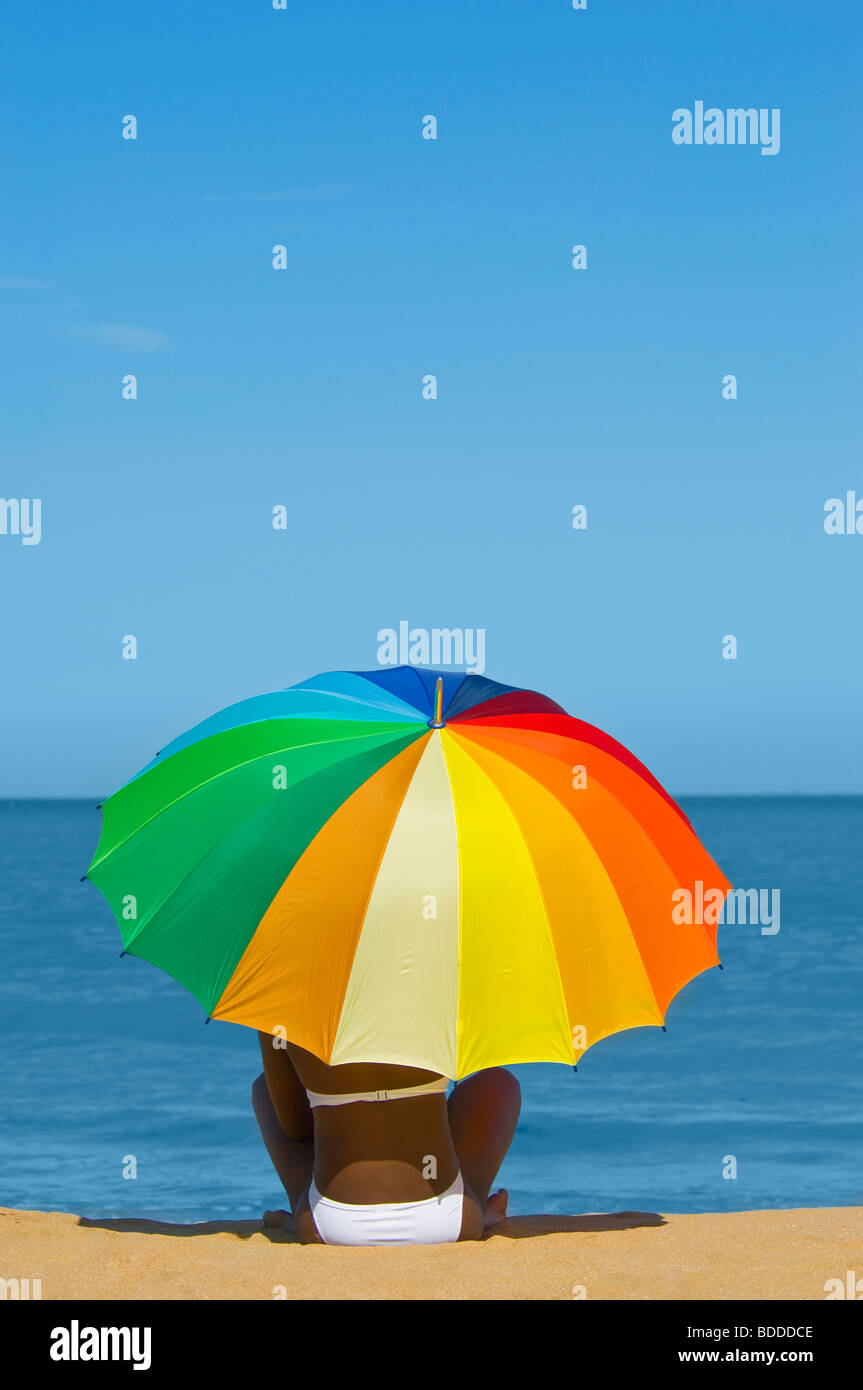 An attractive young African woman holding a multi coloured umbrella sitting on a sandy beach with a blue sea in the background. Stock Photo