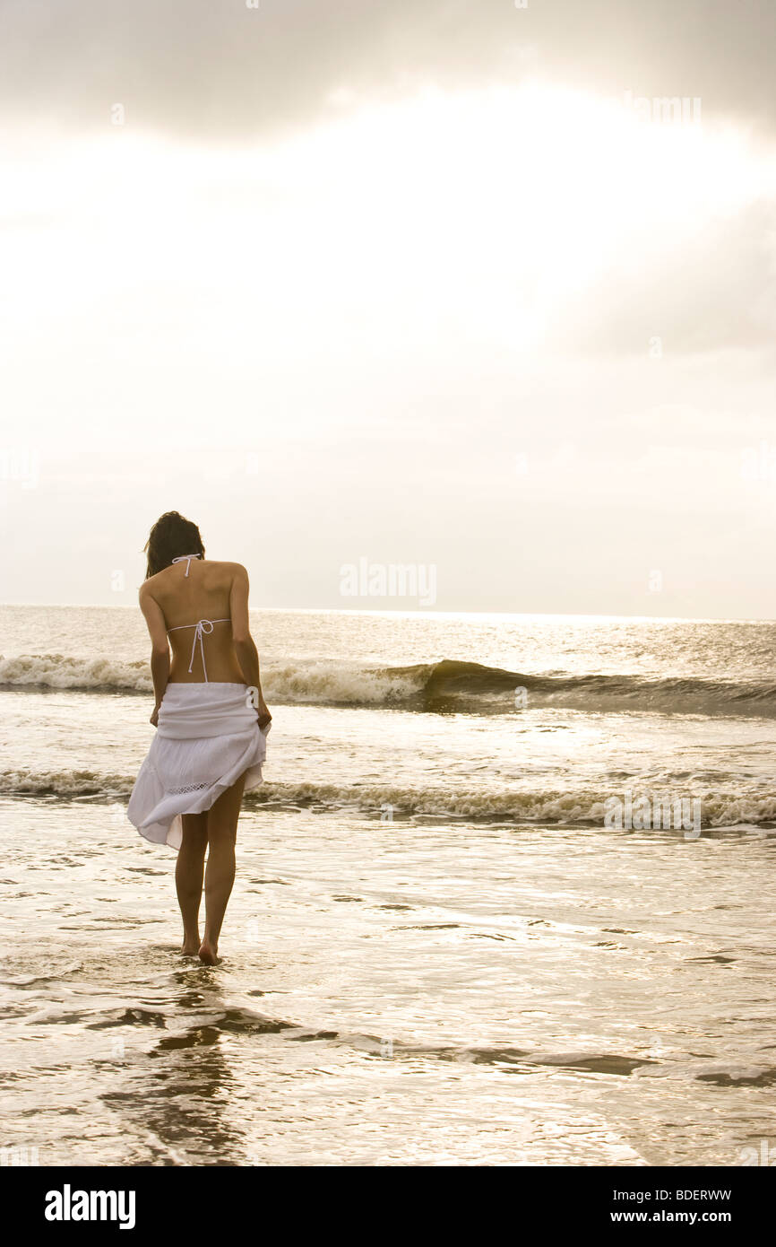 Rear view of young woman in white bikini and skirt walking in ocean at ...