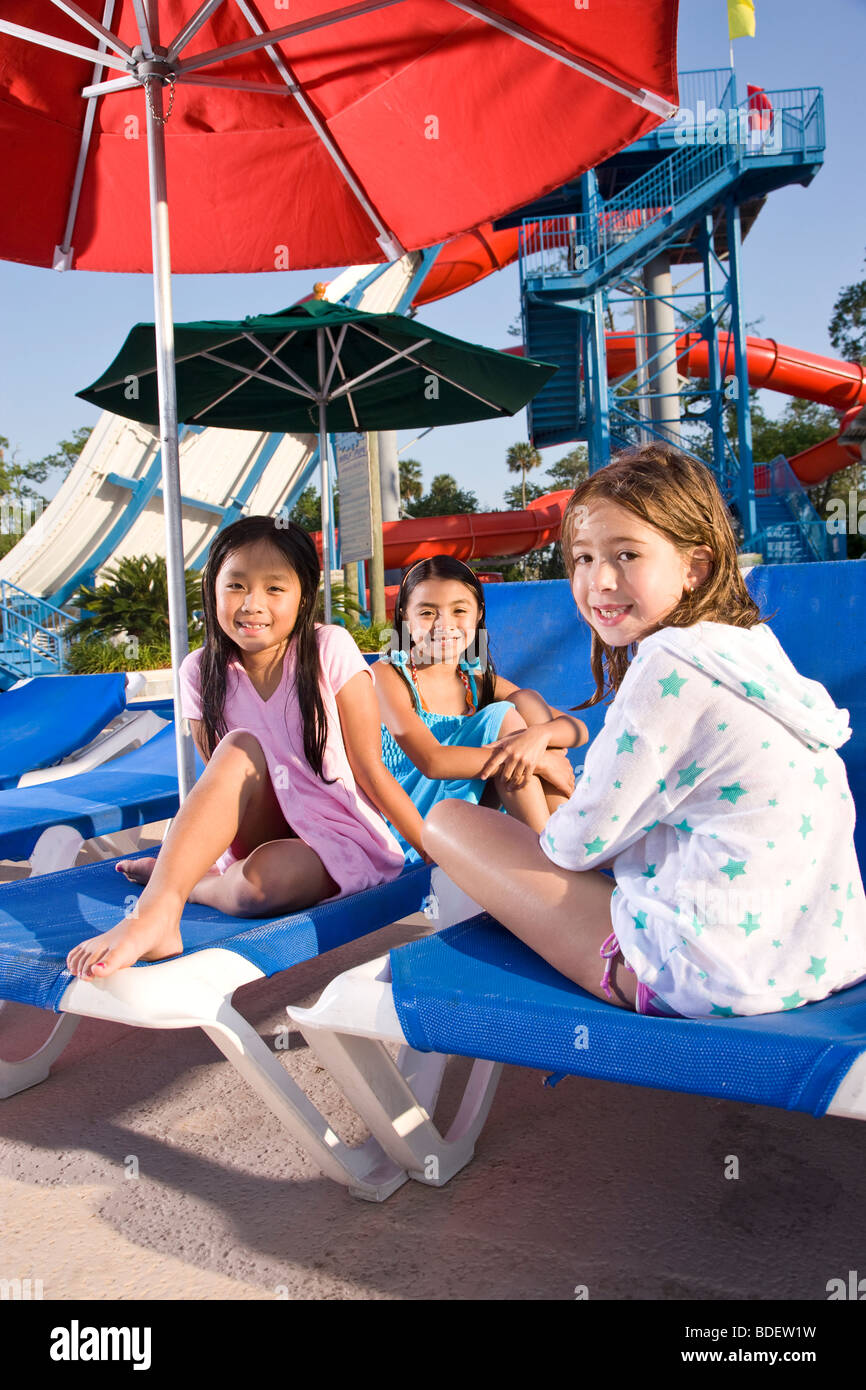 Multi-ethnic girls sitting on deck chairs at water park in summer Stock Photo