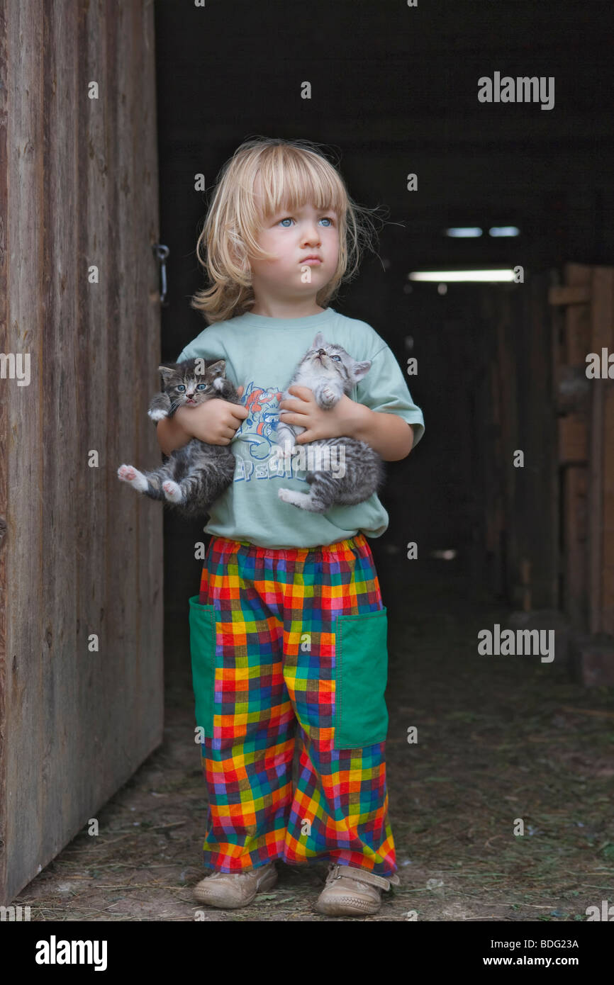 blond boy holding two kittens at the stable door Stock Photo